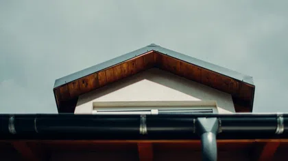 Close-up of a house rooftop with wooden eaves and a black gutter against a cloudy sky.