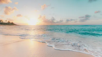 Sunset over a tranquil beach with gentle waves and palm trees on the horizon under a partly cloudy sky.