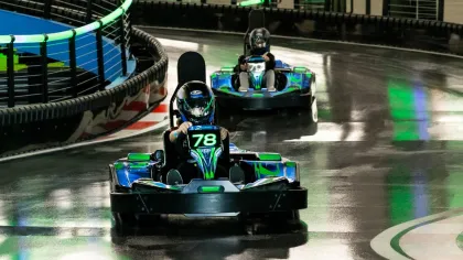 Two racers in helmets driving go-karts on a shiny indoor track with neon green lights and turn arrows.