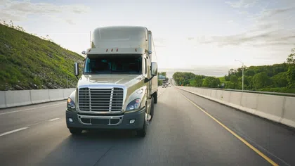Large white semi-truck driving on a highway surrounded by greenery under a bright sky.