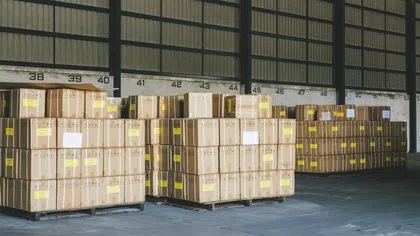 Stacks of sealed cardboard boxes on wooden pallets inside a large warehouse with numbered sections.