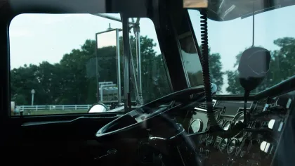 Interior view of a truck cabin featuring the steering wheel, dashboard controls, and side window with an outdoor background.
