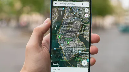 Hand holding smartphone displaying Google Maps satellite view of Manhattan, New York City with landmarks and parks visible.