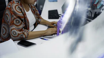 Woman in a colorful shirt typing on a laptop at a white desk with a phone and purple water bottle nearby