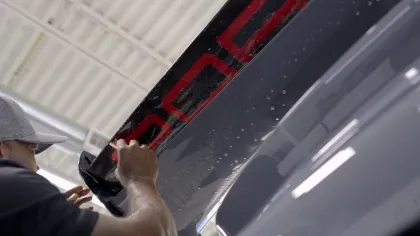 Man installing or adjusting a black spoiler with red lettering on a gray car in a workshop.