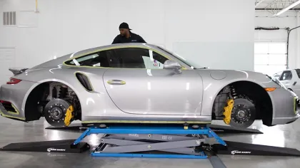 Silver sports car lifted on a blue platform with wheels removed inside a bright garage with a mechanic nearby