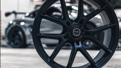 Black multi-spoke car wheel rim in focus with blurred silver sports car in the background inside a garage.