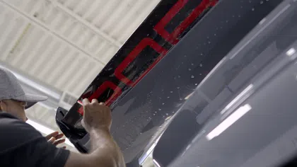 Technician applying black and red decal to a glossy dark vehicle surface inside a workshop with industrial ceiling.
