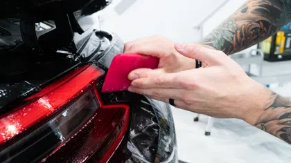 Person applying protective film to a black car's rear taillight using a pink tool in a workshop.