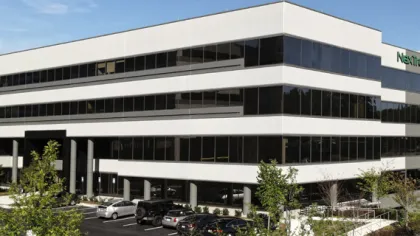 Modern three-story office building with large tinted windows and surrounding parking lot with cars.