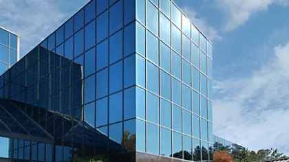 Modern glass office building reflecting blue sky with clouds and surrounding greenery on a sunny day