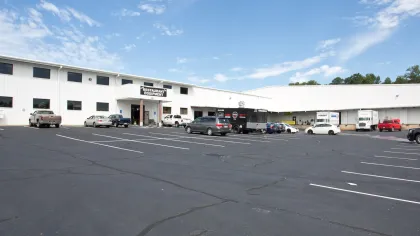 Large white industrial building with multiple parked vehicles and clear blue sky above in spacious parking lot.