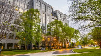 Modern office building with large windows surrounded by green trees and a well-maintained lawn at dusk.