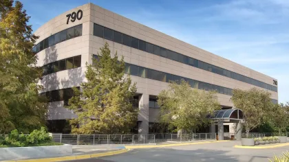 Modern three-story office building numbered 790 surrounded by trees and greenery under a clear sky.