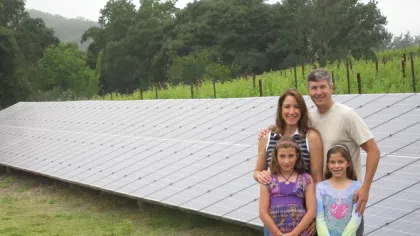 Family of four standing beside a large ground-mounted solar panel array in a grassy rural area with trees behind.