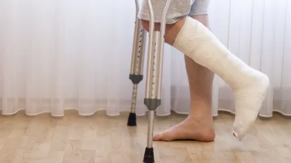 Person with a cast on foot using crutches standing on wooden floor in front of sheer curtains