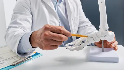 Doctor in white coat pointing at anatomical foot skeleton model with a pencil on white desk.