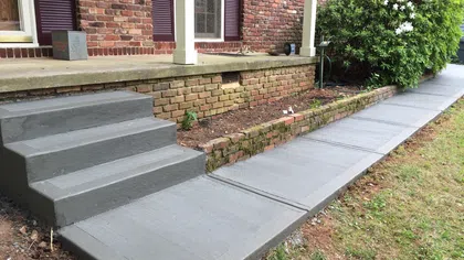Newly poured smooth gray concrete steps and walkway alongside a red brick house with garden area.