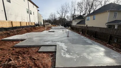Freshly poured concrete foundation slab for a new residential building on red soil between existing houses.