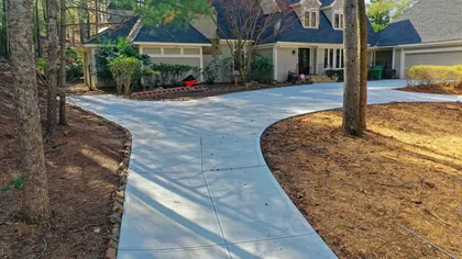 Curved concrete driveway leading to a large suburban house surrounded by trees and landscaped yard.
