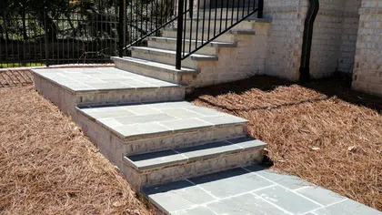 Stone steps with black metal railing leading up to white brick house entrance surrounded by pine straw mulch.
