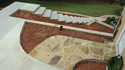 Outdoor stone patio with surrounding bare soil, concrete walkway, and stairway in a garden setting.