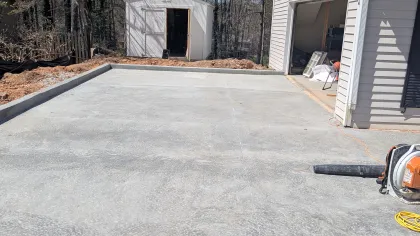Freshly poured concrete driveway next to a white garage and a small white storage shed surrounded by trees.