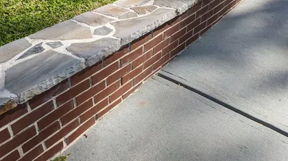 Brick retaining wall with stone cap along concrete sidewalk and green grass lawn in daylight.