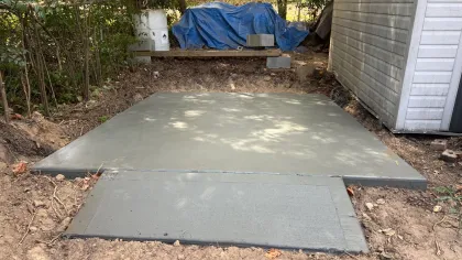 Newly poured smooth concrete slab next to a shed with surrounding dirt and backyard trees.