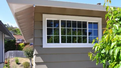 White-framed window on beige siding exterior wall reflecting trees and blue sky outdoors
