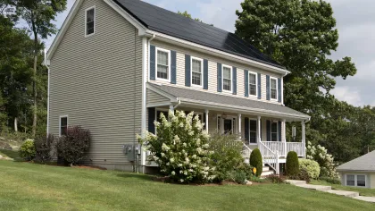 Two-story beige house with solar panels on roof, white porch, and green lawn surrounded by trees and shrubs.