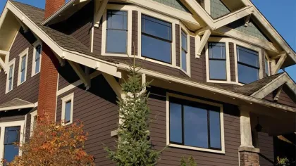 Two-story craftsman-style house with brown siding, white trim, large windows, and a brick chimney under clear blue sky