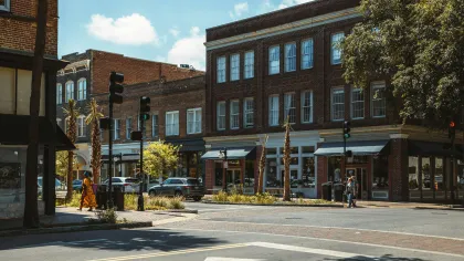 Sunny urban street corner with brick buildings, traffic lights, pedestrians, and cars during the day.