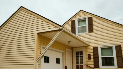 Yellow house with white door, garage, brown shutters, and covered porch under an overcast sky