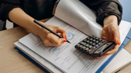 Person with red nail polish using calculator and pen to work on financial documents with stamps.