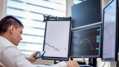 Man analyzing stock market charts on multiple monitors while using a smartphone in a bright office.