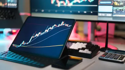 Tablet displaying rising stock market chart with candlesticks and moving averages on a modern trading desk setup.