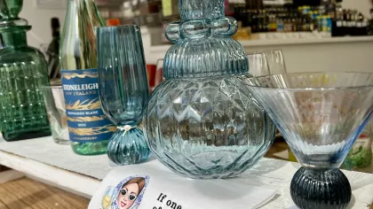 Wine bottle, decorative glassware, and towel with humorous quote about wine on a wooden table in a store.
