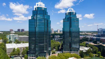 Two modern blue glass skyscrapers with distinct white crown structures surrounded by trees under a bright blue sky.