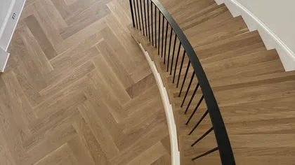 Curved wooden staircase with black railing and chevron-patterned wood flooring in entryway with dark door.