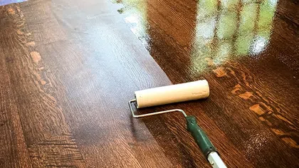 Wood floor being refinished with a paint roller showing wet and dry areas and window reflection.