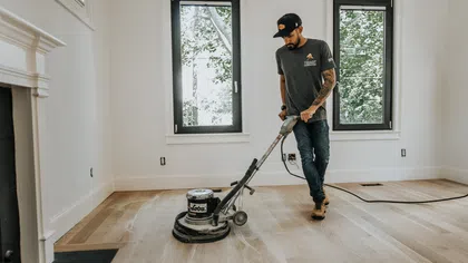 Man refinishing hardwood floor with floor buffer machine in bright room with two windows.