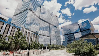 Modern glass office buildings reflecting clouds under a blue sky with trees and urban landscape
