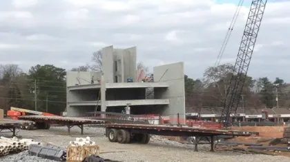 Concrete building under construction with flatbed trailer, crane, and workers on site during daytime.