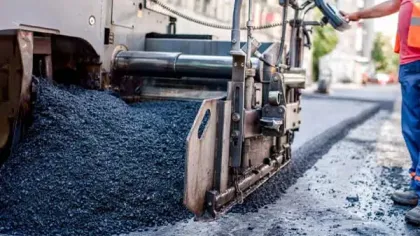 Close-up of asphalt paving machine laying new road surface with worker in safety vest monitoring the process