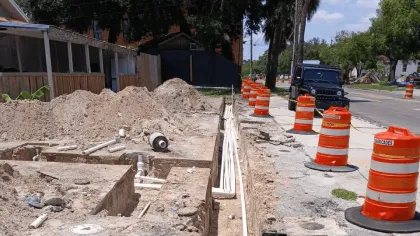 Urban trench construction site with orange traffic barrels, exposed pipes, and dirt piles along a residential street.
