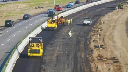 Road construction workers paving a new asphalt lane beside an active highway with traffic moving on existing lanes