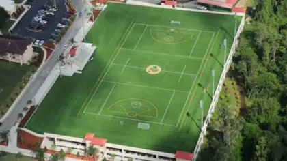 Aerial view of a large green sports field with baseball and soccer markings surrounded by trees and parking areas