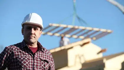 Construction worker in white helmet and plaid shirt stands on site with crane lifting building materials in background.