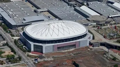 Aerial view of a large oval stadium with a patterned white roof surrounded by industrial buildings and roads.